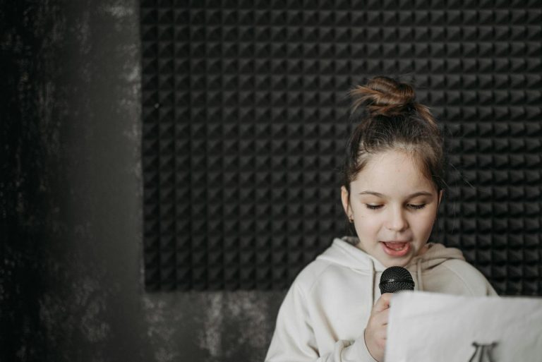 A child with a top bun hairstyle reads from a paper while holding a microphone, standing against a textured wall.