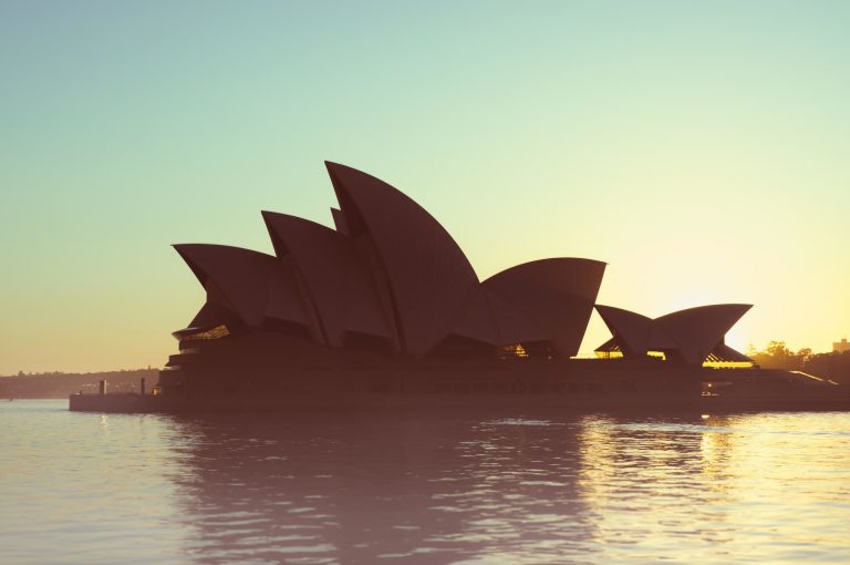 Silhouette of the Sydney Opera House at sunset, with its distinctive sail-like structures reflected in the waters of Sydney Harbour.