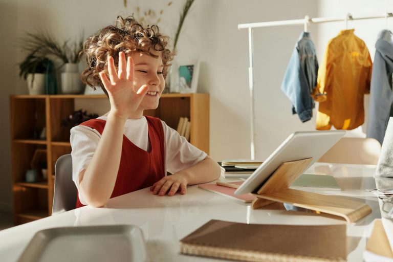 Child with curly hair sitting at a table waves at a tablet placed on a stand. Books and notebooks are on the table, and coats hang in the background.