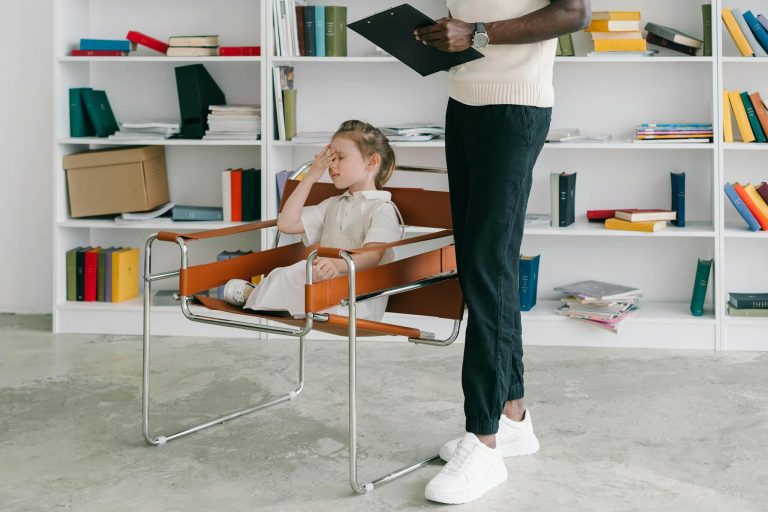 A child sits in a leather chair with a hand on their forehead, while an adult stands nearby holding a clipboard. Books and papers are on white shelves in the background.