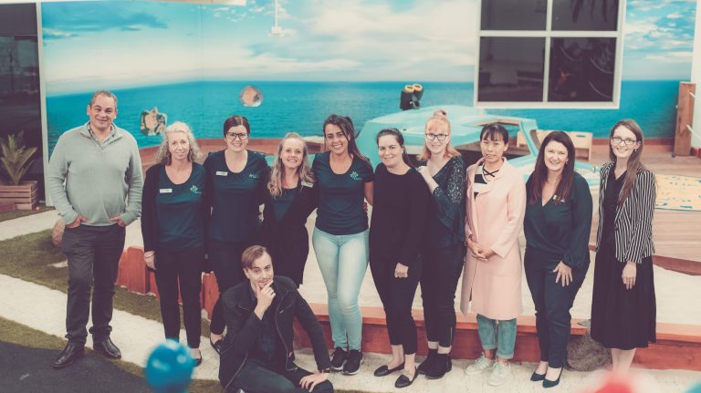 A group of eleven people, some seated and some standing, pose for a photo indoors in front of a beach-themed backdrop.