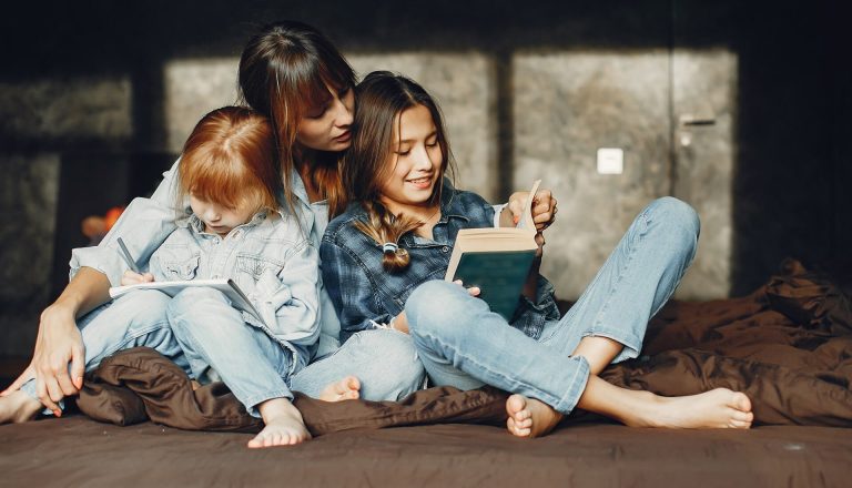 A woman sitting with two children, one reading a book and the other drawing. They are all dressed in denim clothing and seated on a bed.