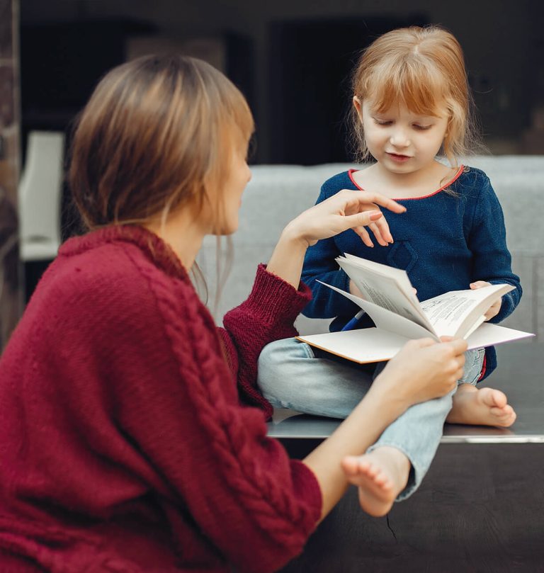 An adult in a red sweater reads a book to a young child sitting on a table, both engaged in the activity.