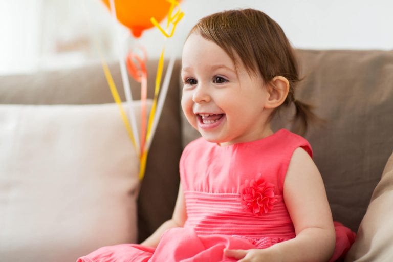 A smiling toddler in a pink dress sits on a beige couch with orange balloons in the background, celebrating World Autism Awareness Day 2019.