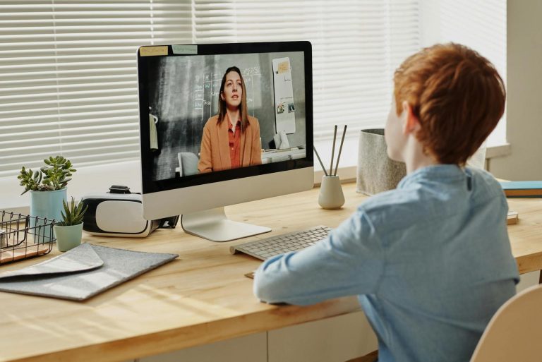 A person with red hair sits at a desk, watching a videocall on a computer.