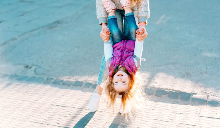 Person holding a smiling child upside down by the hands on a sunny day. The child is wearing jeans, a pink vest, and sneakers.