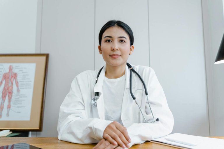 A doctor in a white coat with a stethoscope sits at a desk with medical charts and a lamp nearby.