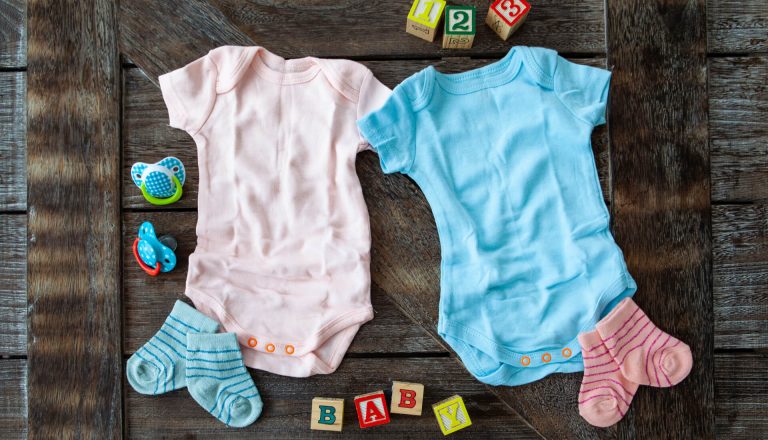 Two baby onesies, one pink and one blue, are laid out on a wooden surface with matching socks and various colorful toys and alphabet blocks around them.