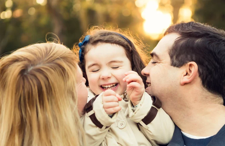 A child laughs joyfully between two adults, likely parents, outdoors with sunlight in the background, creating a moment of pure happiness and awareness for the beauty of everyday life.