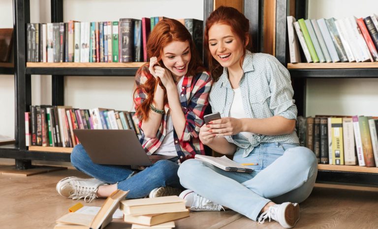 Two women sitting on the floor in a library, looking at a smartphone. One has a laptop and notebook, and there are several books around them.