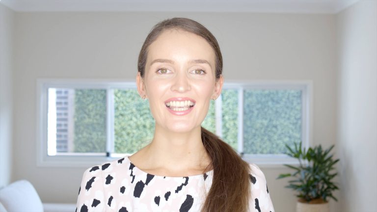 A woman with long brown hair, wearing a black and white patterned blouse, smiles in a brightly lit room with green foliage visible through large windows behind her.