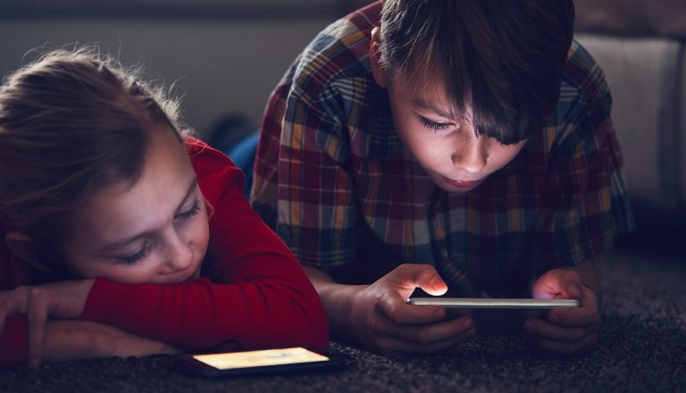 Two children lie on the floor, each focused on handheld electronic devices. One child wears a red shirt and the other wears a plaid shirt.