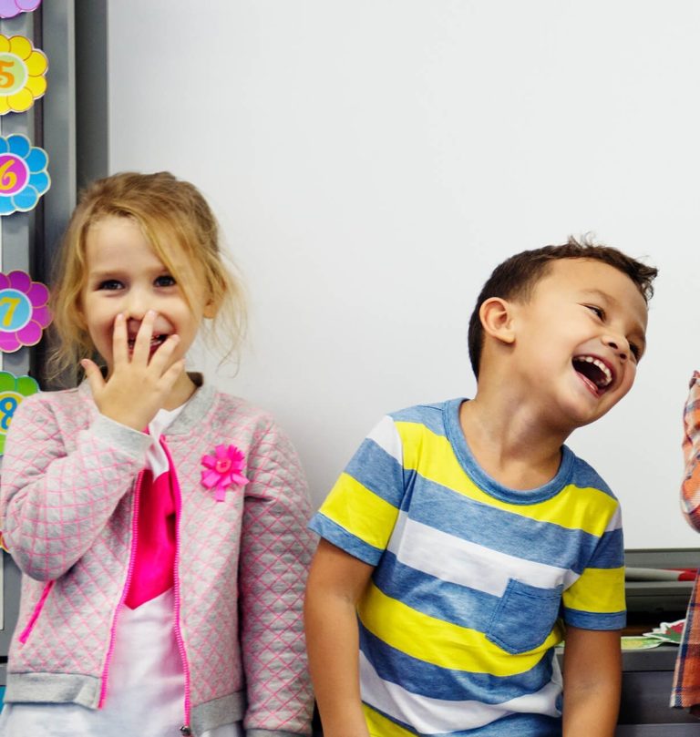 A young girl and boy stand in a classroom; the boy is laughing while the girl covers her mouth with her hand. Colorful decorations are visible in the background.