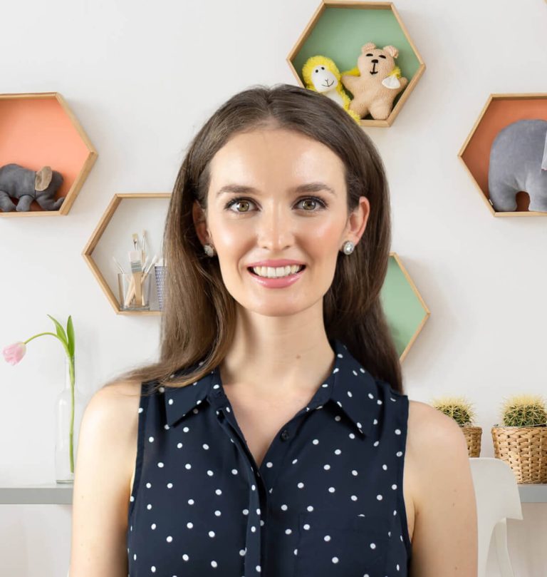 A woman with long brown hair and a polka dot sleeveless blouse smiles in front of hexagonal shelves with toys and decorations.