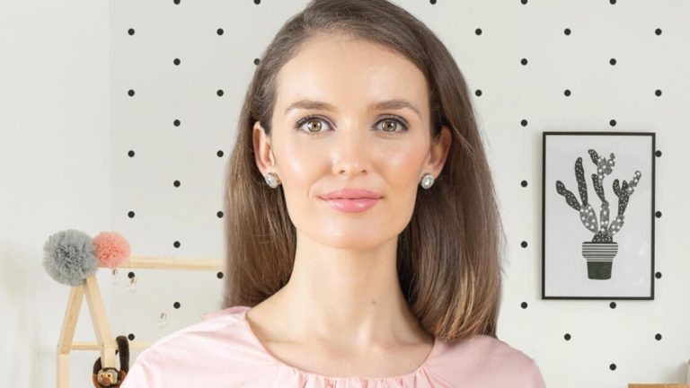 A woman with shoulder-length brown hair, wearing a light pink blouse, stands in a room with polka dot wallpaper and a framed cactus print on the wall.