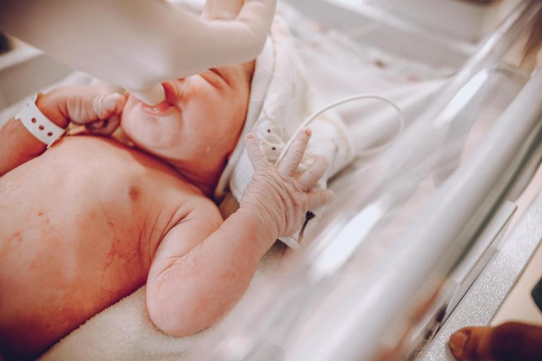 Newborn baby in a hospital crib, wearing a wristband, receiving care from a gloved hand.