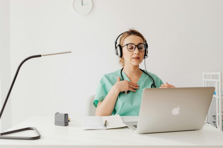 A healthcare professional in scrubs and headphones speaks during a video call, sitting at a desk with a laptop, notebook, and pen.