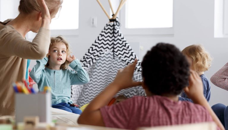 A teacher and children engage in an activity in a classroom with a play tent and toy blocks in the background.