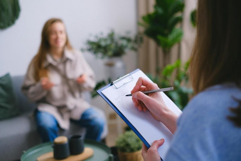 Person taking notes on a clipboard while talking to another person sitting on a sofa.