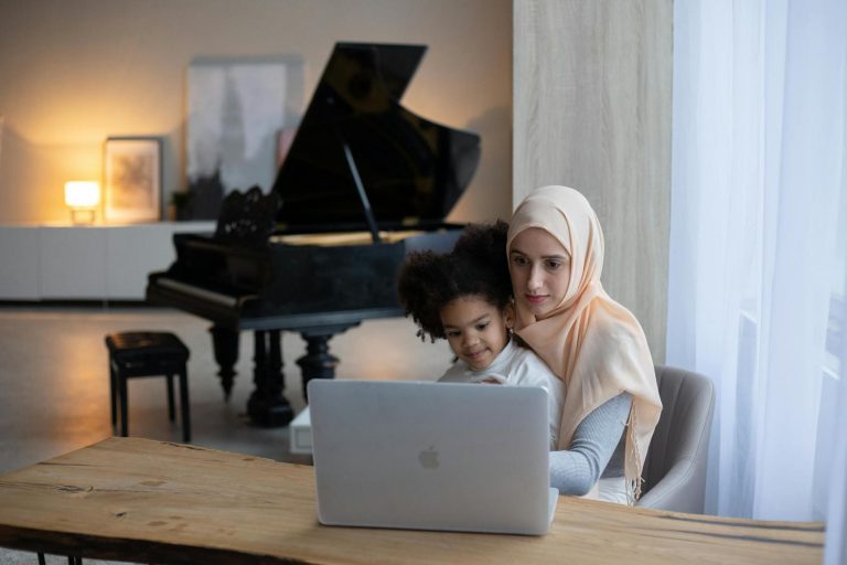 A woman and a child sit at a wooden table, using a laptop. A grand piano is visible in the softly lit background.