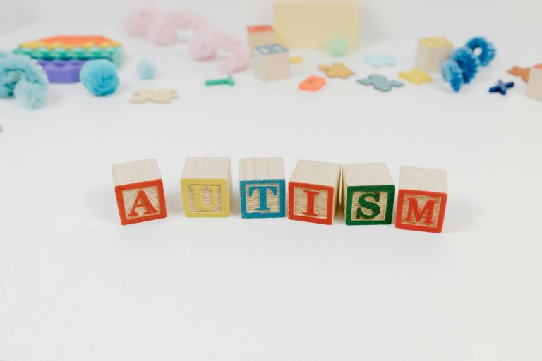 Wooden blocks spell "AUTISM" on a white surface, surrounded by colorful toys and shapes in the background.