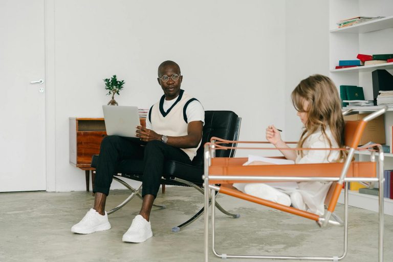 An adult and a child sit in a minimalist room. The adult is on a laptop, and the child is drawing. White walls and shelves with books are in the background.