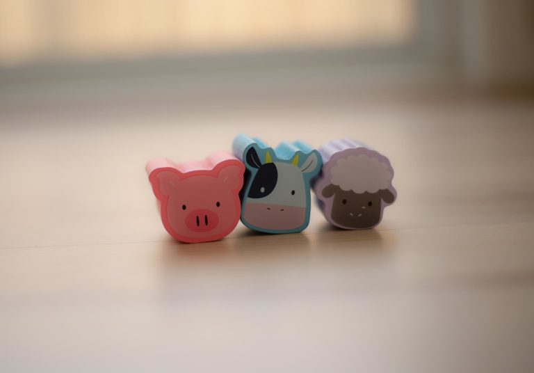 Three cute, colorful plastic animal toys (a pig, a cow, and a sheep) are lined up in a row on a wooden floor.