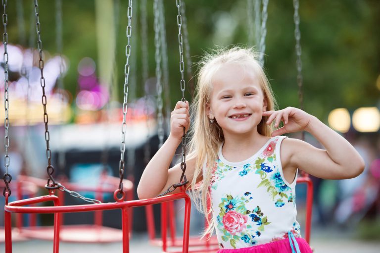 A young girl with blonde hair, wearing a floral top and pink skirt, smiles while standing next to an empty carnival swing ride.