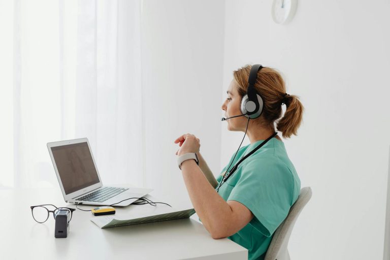 A healthcare professional in scrubs and headset sits at a desk, using a laptop for a video call. Glasses and medical equipment are nearby.